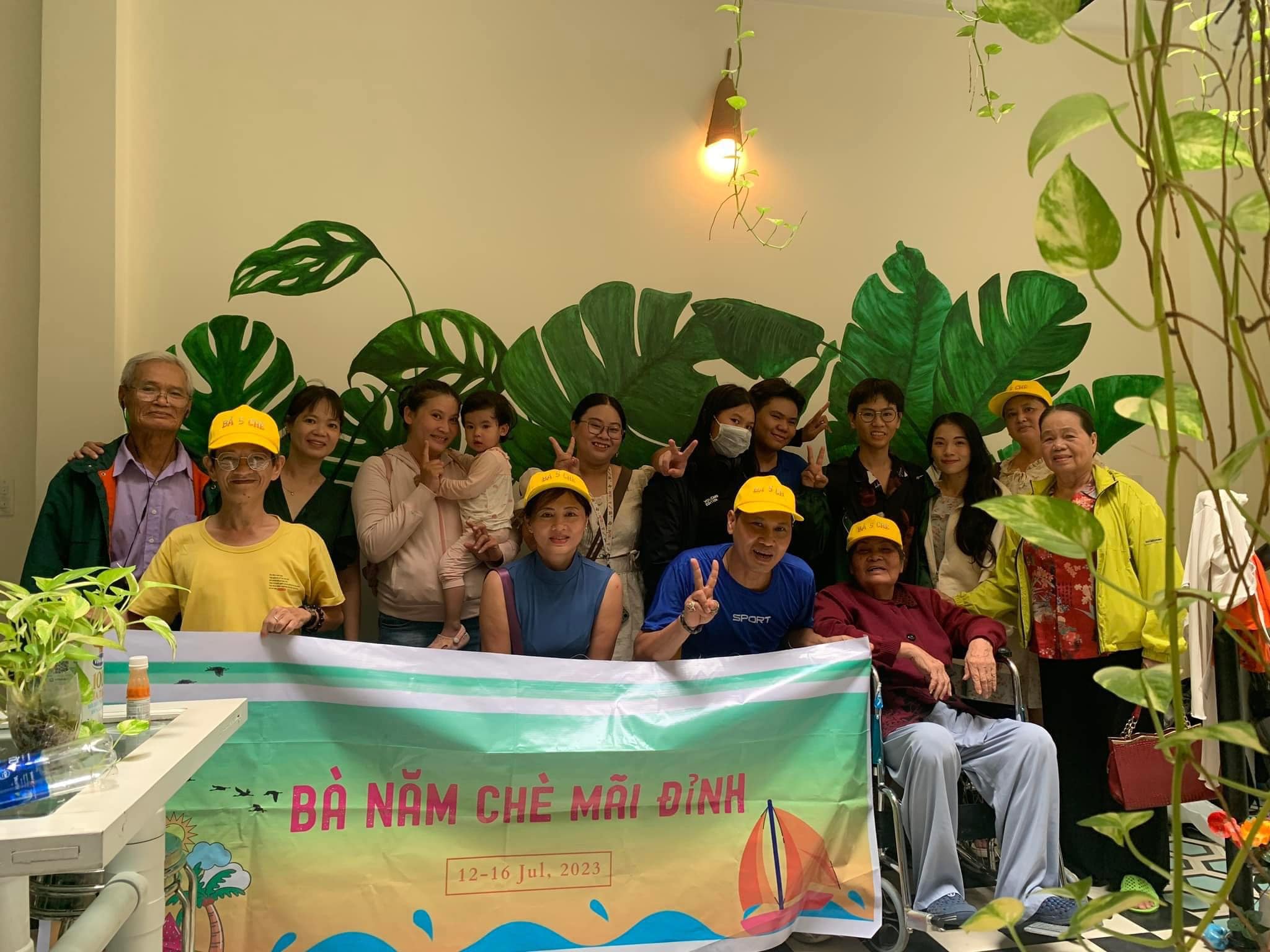 Gia đình nhiều thế hệ chụp ảnh kỷ niệm cùng nhau trong không gian homestay tại Huế, cầm banner chuyến đi, phía sau là tường trang trí lá xanh.