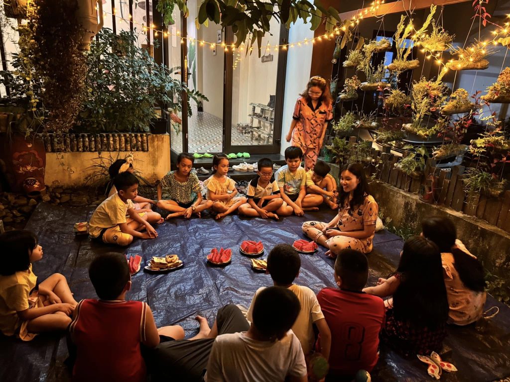 Nhóm trẻ em ngồi thành vòng tròn trong sân vườn homestay tại Huế vào buổi tối, cùng ăn dưa hấu và bánh dưới ánh đèn dây và cây xanh.