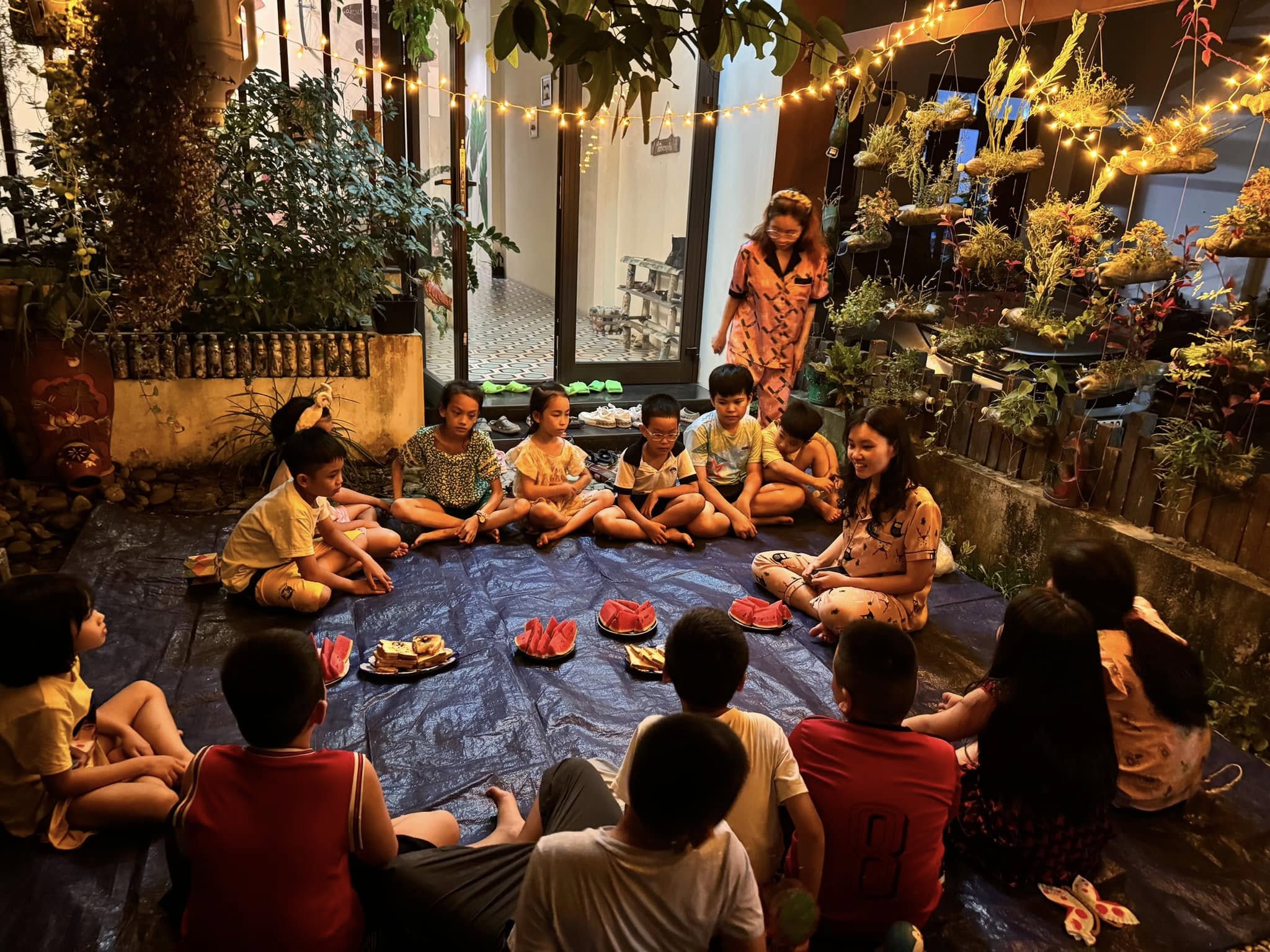 Nhóm trẻ em ngồi thành vòng tròn trong sân vườn homestay tại Huế vào buổi tối, cùng ăn dưa hấu và bánh dưới ánh đèn dây và cây xanh.